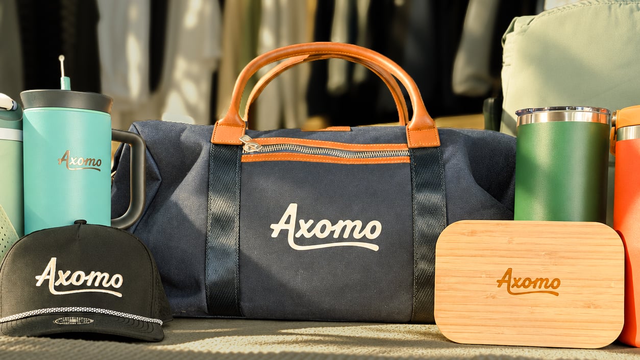 A hat, a bag, a wooden lunch box and a tumbler with the logo Axomo on each of them